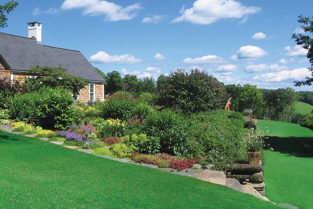 Expansive lawns surround a garden lined with a stonewall on a slope next to a house.