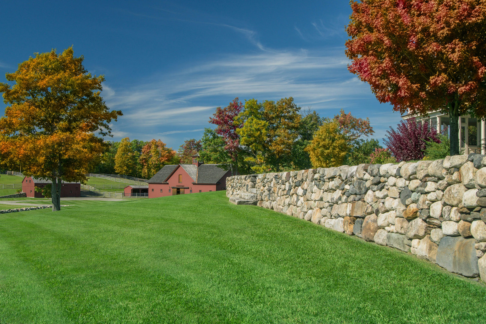 Healthy green grass lawn bordered by fall trees, a stonewall and red barn and outbuildings.